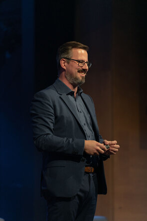 Ein l&auml;chelnder Mann in einem blauen Anzug steht auf einer B&uuml;hne und h&auml;lt ein Smartphone in der Hand.