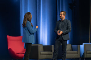 Zwei Personen im Gespr&auml;ch auf einer B&uuml;hne mit blauen Vorh&auml;ngen