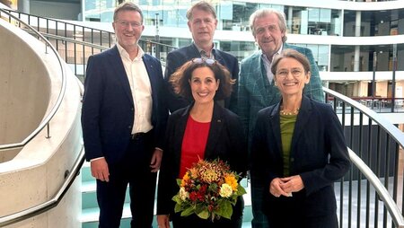 Fünf Personen stehen auf einer Treppe in einem modernen Gebäude, lächelnd und in formeller Kleidung, während eine Frau einen Blumenstrauß hält.