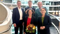 Fünf Personen stehen auf einer Treppe in einem modernen Gebäude, lächelnd und in formeller Kleidung, während eine Frau einen Blumenstrauß hält.