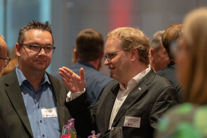 Zwei M&auml;nner unterhalten sich angeregt auf einer Veranstaltung, w&auml;hrend im Hintergrund weitere G&auml;ste zu sehen sind.