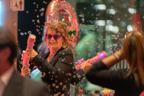 Eine fr&ouml;hliche Frau mit einer Leopardenm&uuml;tze und Sonnenbrille spr&uuml;ht Seifenblasen mit einer pinken Wasserpistole.