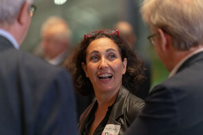 Eine Frau mit lockigem Haar und roten Brillen sitzt in einer Gruppe und l&auml;chelt, w&auml;hrend sie mit anderen spricht.