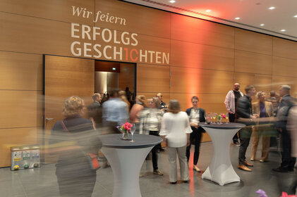 Eine lebhafte Veranstaltung mit Gästen, die an Stehtischen stehen und sich unterhalten, während ein Banner im Hintergrund sichtbar ist.