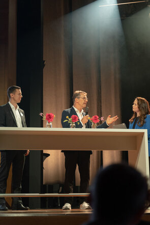 Drei Personen stehen an einem Tisch, w&auml;hrend einer von ihnen mit einem Mikrofon spricht und die anderen aufmerksam zuh&ouml;ren.