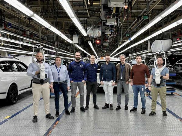 Eine Gruppe von acht M&auml;nnern steht in einer modernen Autofabrik, umgeben von wei&szlig;en Fahrzeugen und industrieller Ausr&uuml;stung.