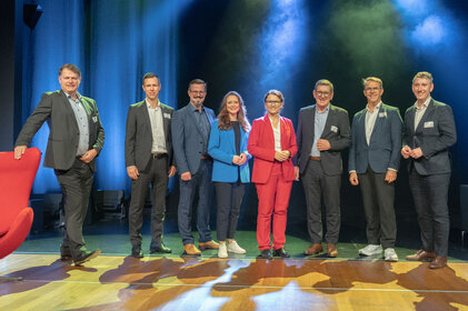 Eine Gruppe von acht Personen steht auf einer Bühne, umgeben von buntem Licht, und lächelt in die Kamera.