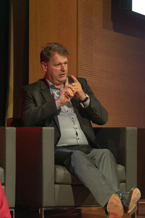 Ein Mann in einem Anzug sitzt auf einem Stuhl und gestikuliert mit seinen H&auml;nden, w&auml;hrend er spricht.