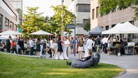 Eine lebhafte Gartenparty mit Gästen, die an Stehtischen stehen und sich unterhalten, umgeben von Bäumen und Zelten.