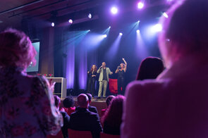 Drei S&auml;nger stehen auf der B&uuml;hne, w&auml;hrend das Publikum begeistert applaudiert und die Lichter in bunten Farben leuchten.