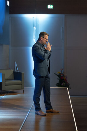 Ein Mann in einem Anzug steht auf einer B&uuml;hne und h&auml;lt seine H&auml;nde in einer nachdenklichen Geste, umgeben von sanftem Licht.