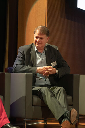 Ein Mann in einem Anzug sitzt entspannt auf einem Sessel und l&auml;chelt, w&auml;hrend er an einer Diskussion teilnimmt.