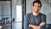 Ein junger Mann mit einem freundlichen Lächeln steht in einem modernen Büro und hat die Arme vor der Brust verschränkt.