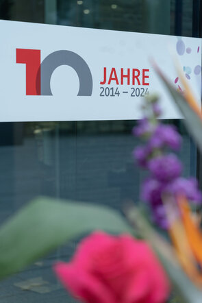 Ein Banner feiert 10 Jahre von 2014 bis 2024, umgeben von bunten Blumen und einer modernen Glasarchitektur.