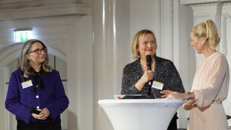 Drei Frauen stehen an einem Podium, während eine von ihnen spricht und die anderen aufmerksam zuhören.