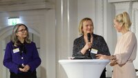 Drei Frauen stehen an einem Podium, während eine von ihnen spricht und die anderen aufmerksam zuhören.