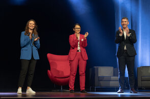 Drei Personen stehen auf einer B&uuml;hne und applaudieren, w&auml;hrend ein roter Sessel im Hintergrund sichtbar ist.