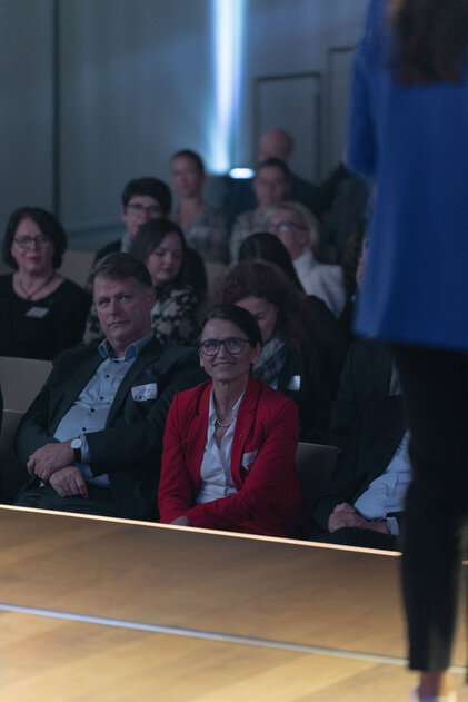 Eine Gruppe von Menschen sitzt aufmerksam im Publikum, während eine Person auf der Bühne spricht. Die Atmosphäre ist konzentriert.