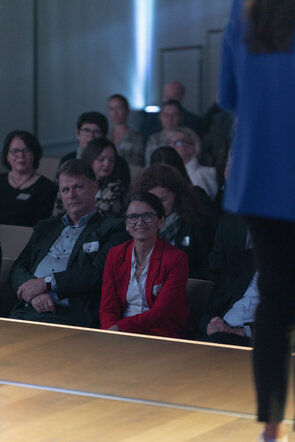 Eine Gruppe von Menschen sitzt aufmerksam im Publikum, w&auml;hrend eine Person auf der B&uuml;hne spricht. Die Atmosph&auml;re ist konzentriert.