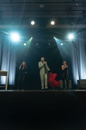 Drei S&auml;nger stehen auf einer B&uuml;hne, umgeben von bunten Lichtern, w&auml;hrend sie leidenschaftlich in Mikrofone singen.