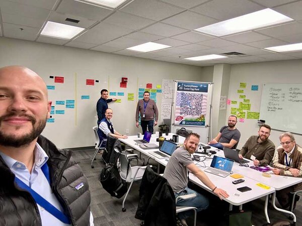 Eine Gruppe von f&uuml;nf M&auml;nnern sitzt an einem Tisch, w&auml;hrend ein weiterer Mann an einer Wand mit Notizen arbeitet.