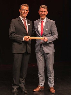 Zwei M&auml;nner &uuml;berreichen sich eine Auszeichnung auf der B&uuml;hne