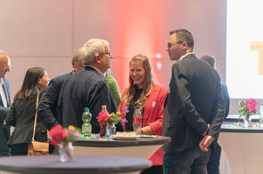 Eine Gruppe von Fachleuten unterh&auml;lt sich lebhaft bei einer Veranstaltung, w&auml;hrend bunte Blumen auf den Tischen stehen.