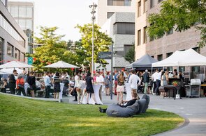 Eine lebhafte Gartenparty mit G&auml;sten, die an Stehtischen stehen und sich unterhalten, umgeben von B&auml;umen und Sonnenschirmen.