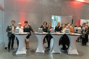 Eine Gruppe von f&uuml;nf Frauen und einem Mann steht an Empfangstischen, die mit Blumen und Registrierungsunterlagen dekoriert sind.