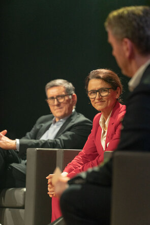 Drei Personen sitzen auf einer B&uuml;hne, w&auml;hrend sie sich angeregt unterhalten, mit einem Fokus auf die Frau in einem roten Blazer.