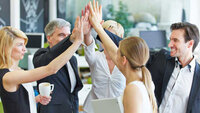 Eine Gruppe von fünf Personen in einem modernen Büro gibt sich gegenseitig High-Fives, während sie lächeln und Freude ausstrahlen.