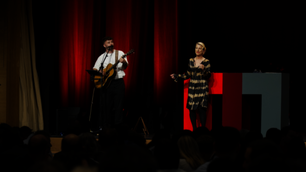 Ein Musiker mit Gitarre steht auf der B&uuml;hne, w&auml;hrend eine S&auml;ngerin in einem gestreiften Kleid das Publikum begeistert.