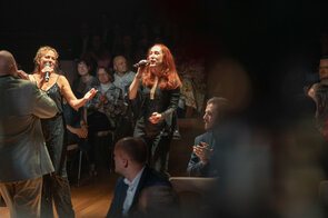 Zwei S&auml;ngerinnen in glitzernden Outfits stehen auf der B&uuml;hne und singen leidenschaftlich ins Mikrofon, w&auml;hrend das Publikum applaudiert.