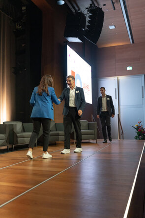Eine Frau in einem blauen Blazer sch&uuml;ttelt einem Mann die Hand, w&auml;hrend ein anderer Mann im Hintergrund auf die B&uuml;hne geht.