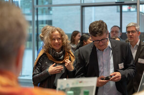 Eine fr&ouml;hliche Frau mit lockigem Haar l&auml;chelt, w&auml;hrend sie sich in einer Gruppe von Menschen in einem modernen Raum befindet.