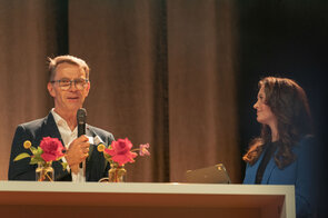 Ein l&auml;chelnder Mann mit Brille h&auml;lt ein Mikrofon, w&auml;hrend eine Frau in einem blauen Blazer ihm aufmerksam zuh&ouml;rt.