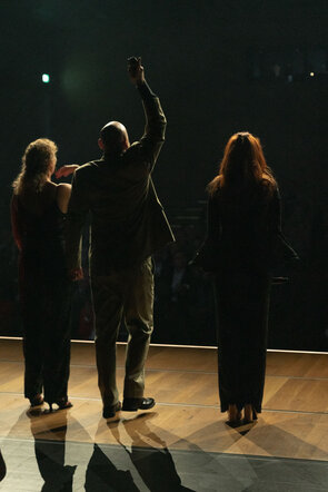 Drei Personen stehen auf einer B&uuml;hne, w&auml;hrend sie dem Publikum zuwinken, umgeben von schwachem Licht und Schatten.