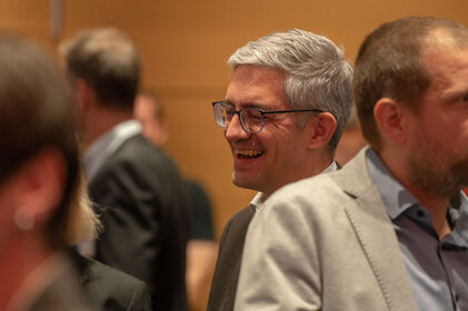 Ein Mann mit grauen Haaren und Brille lächelt fröhlich in einer Gruppe von Menschen in einem Konferenzraum.