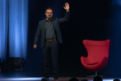 Ein Mann in einem Anzug hebt die Hand, während er auf einer Bühne steht, mit einem roten Stuhl im Hintergrund.